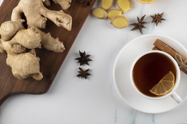 Taza de té con rama de canela y un trozo de limón, cerca de una tabla con una raiz de jengibre. 